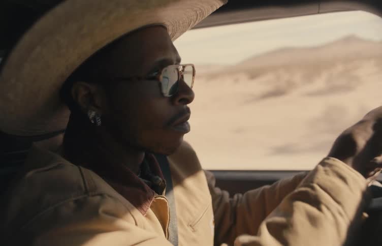A man in a cowboy hat looks around as he drives through an arid landscape