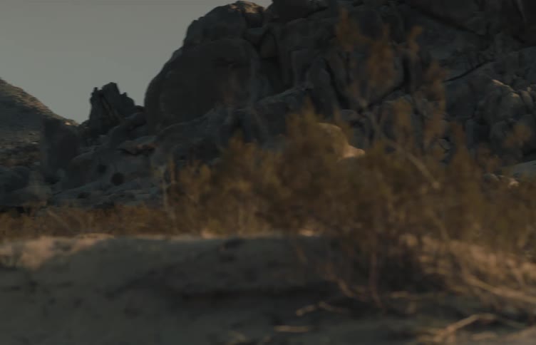 A dark offroad vehicle maneuvers across a rugged desert landscape filled with large rock formations and sparse vegetation the camera pans as the vehicle navigates the sandy terrain during the golden hour eventually disappearing behind dunes and brush