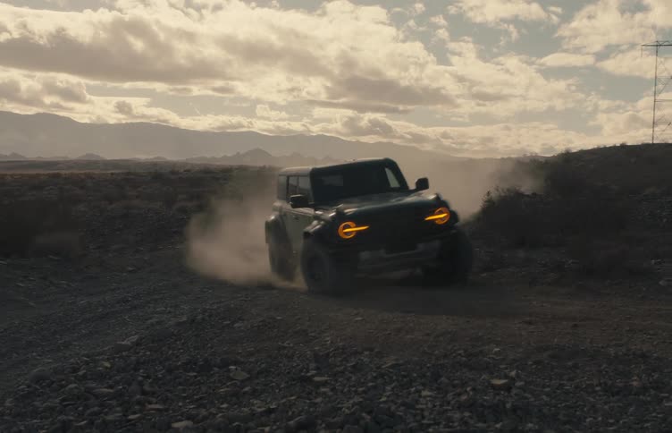 A dark offroad vehicle with glowing orange headlights speeds across a dusty desert dirt road the car kicks up a thick cloud of dust as it passes a power line tower under a cloudy lateafternoon sky