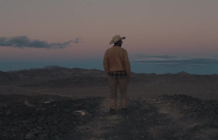 Man wearing a hat standing on top of a hill observes mountains at dusk
