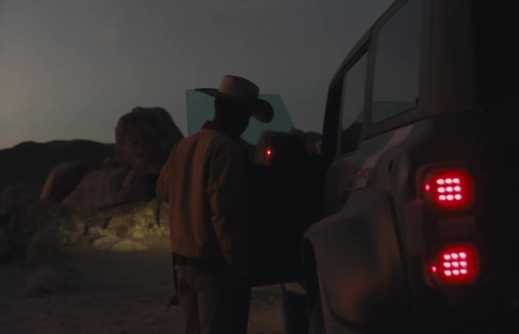 Tail lights glow on a jeep parked in the desert as a man walks and opens the door to get inside