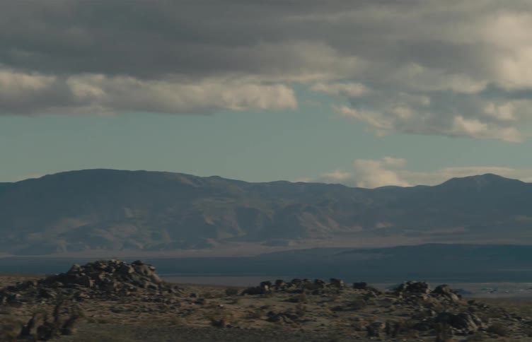 The camera pans across a vast desert landscape featuring rugged dark mountains and rolling hills under a cloudy sky while sparse shrubs and rocky patches cover the foreground of the arid valley floor during the day