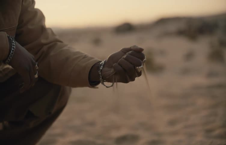 A man wearing a cowboy hat and tan jacket kneels in a desert at dusk and slowly lets fine sand pour through his fingers from a closed fist until his palm is empty against the blurred horizon