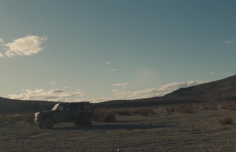 A lone suv sits parked in a vast desert landscape under a wide blue sky as a man standing a few feet behind it walks up and gets inside