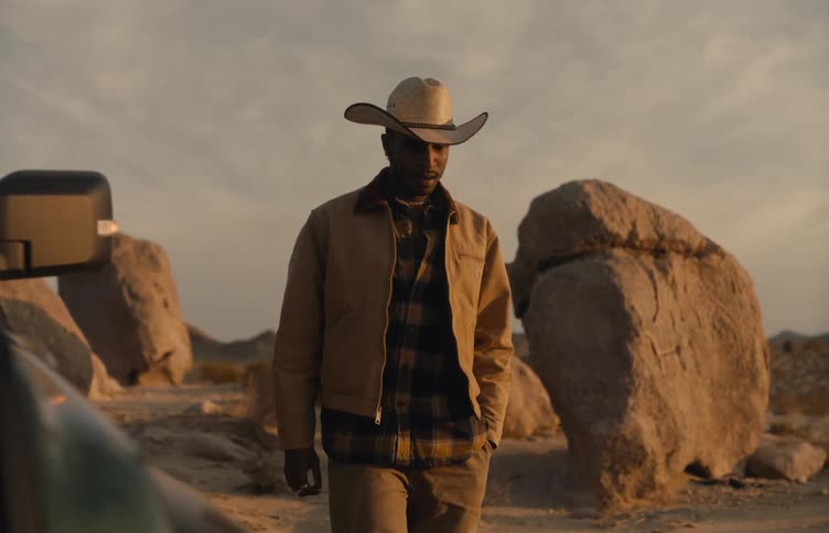 A man wearing a tan jacket and a cowboy hat gets out of a dark vehicle and stands in a desert during a bright sunset with large rocks and mountains in the background