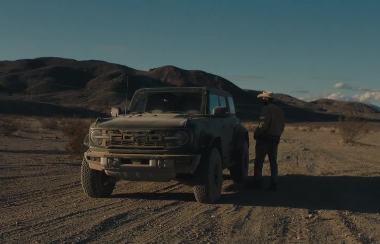 A young man wearing a brown jacket and a cowboy hat walks through the wilderness toward his ford suv covered in dust as he gets inside