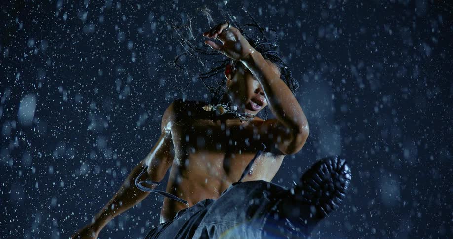 Shirtless man performs a movement under heavy rain with blue lighting and visible droplets