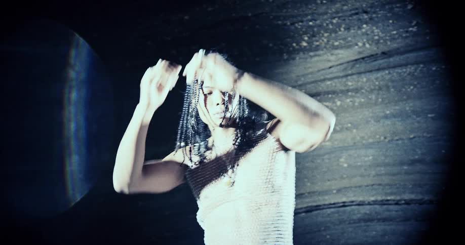 A man with long braids and a white mesh shirt poses with his hands on his head in a studio setting where dramatic light creates striped shadows across his face and body