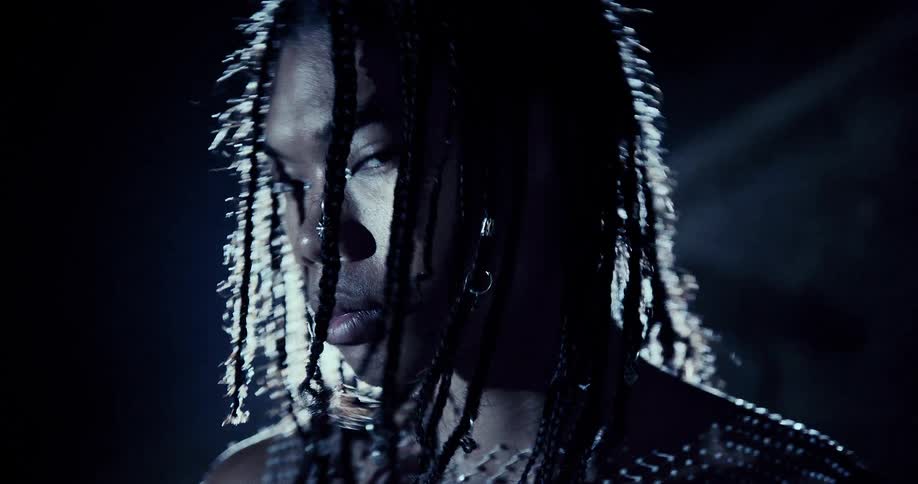 A woman with braided hair stands in moody blue lighting her face turned to the side as light catches the texture of her clothing and jewelry