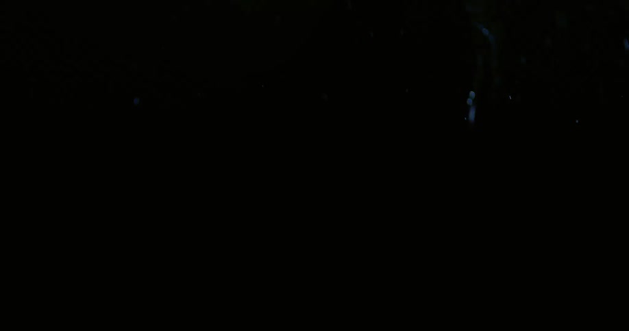 A person with braided hair and face jewelry throws a punch outdoors at night while bright lights shine and create a blue glow against the dark sky and rocky terrain