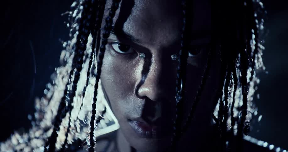 A person with braided hair and face jewelry stands outdoors at night while bright lights shine and create a blue glow against the dark sky and rocky terrain