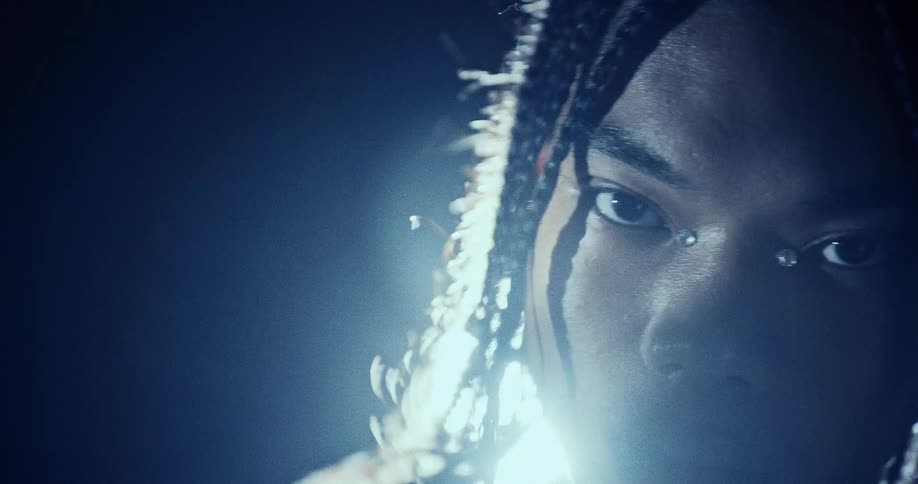 A close up of a black man with long braids and silver jewelry under his eyes as he stares intensely into the camera while bright blue light creates heavy flares and shadows across his face in a dark setting