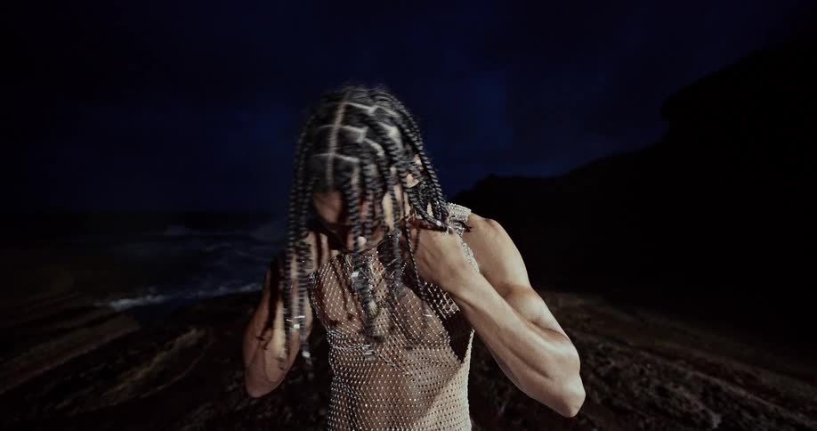 Man wearing a metal mesh shirt stands near the coast at night