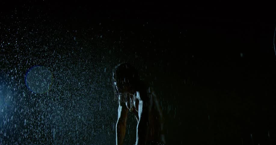 A person stands in front of a large screen in what appears to be a studio setting drenched as water pours down dramatically in the dark