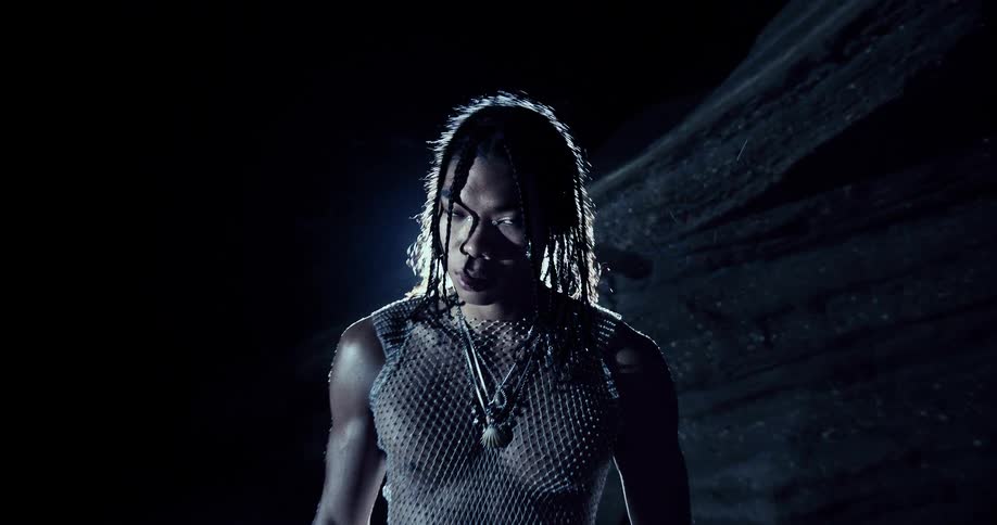 A person with braided hair and face jewelry stands outdoors at night while bright lights shine and create a blue glow against the dark sky