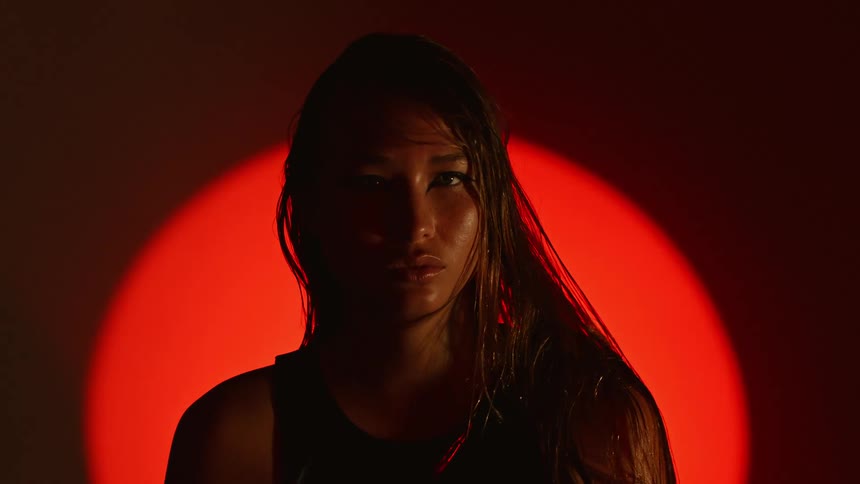 A woman with damp hair and a focused expression gazes directly at the camera in a moody lowlight settinga vibrant orange circular light creates a bold silhouette behind her highlighting her features against the dark background