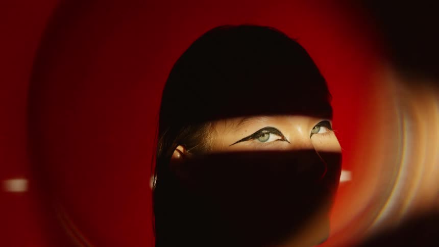 Band of light reveals the eyes of a woman wearing makeup in a shadowed area with a red pattern in the background