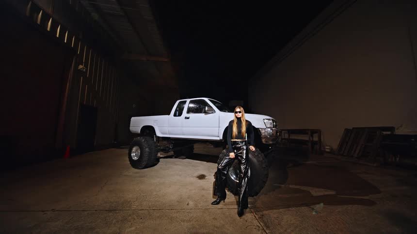A woman in edgy black attire and sunglasses poses confidently against the massive tire of a lifted white pickup truck at nightthe industrial setting and dramatic lighting emphasize the sharp contrast between the sleek fashion and the rugged oversized
