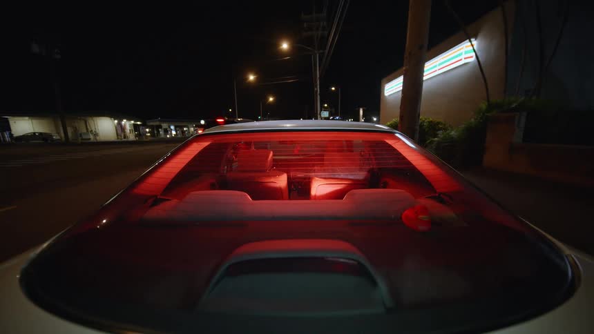 A view from behind a white car shows red lights glowing through the rear window while the vehicle sits on a street at night