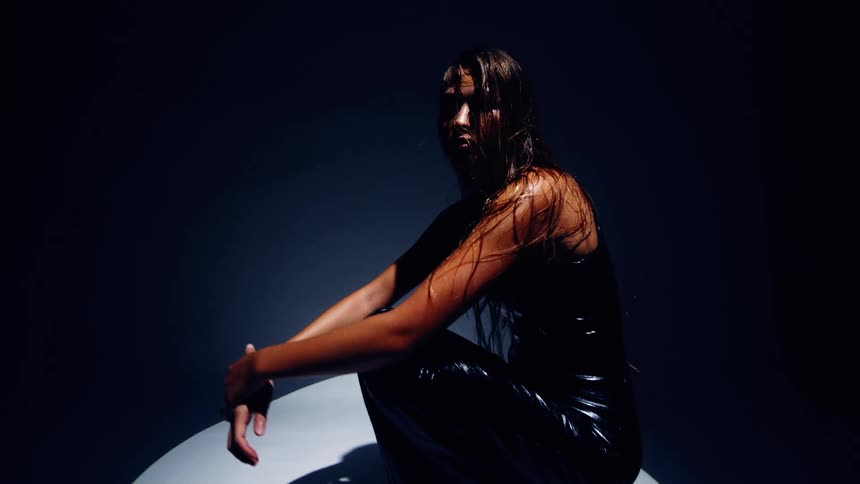 A woman with wet hair and a glossy black outfit sits on a white platform under dramatic blue lighting looking forward