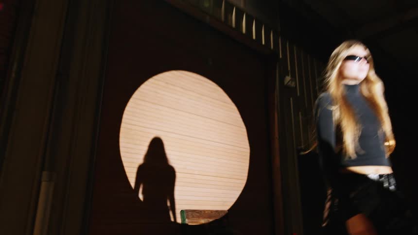The video shows a woman with long hair walking past a circular light projection on a building at nightshe wears dark clothing and sunglasses moving from the shadows into the light as she passes a white vehicle