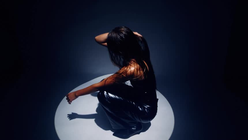 A person in dark clothing crouches in a spotlight on a dark stage