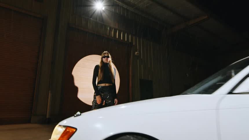 A woman wearing sunglasses and dark clothing stands next to two vehicles in an industrial area at night with rain falling