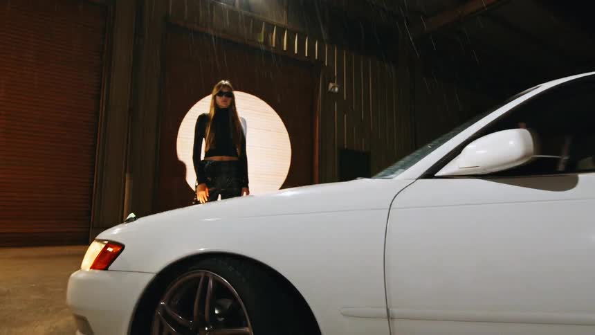 A woman with long blonde hair and sunglasses stands outside at night next to a white car while rain falls and a bright light shines from a circular opening in a building behind her