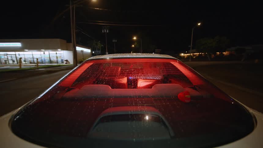 A camera in a back of a white car looks through the rear window at a dark street as the vehicle travels past street lights and buildings during the night