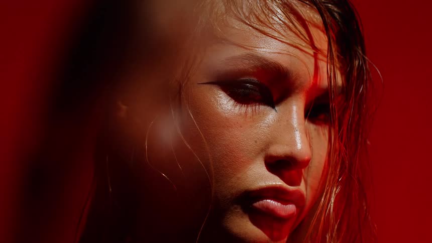 A closeup portrait of a woman with wet hair and dramatic eye makeup illuminated by intense red lighting that casts bold shadows across her face