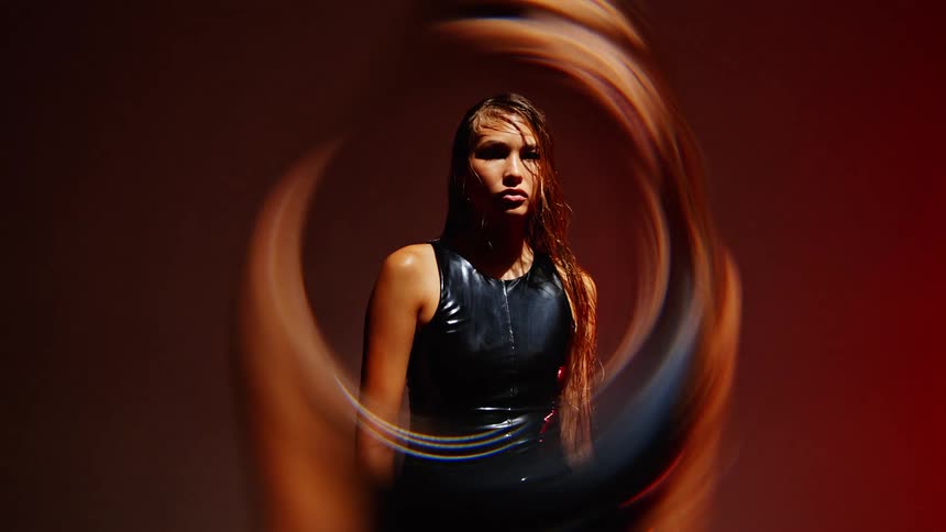 A woman with long wet hair wearing a black outfit is seen through a circular lens against a shifting red background