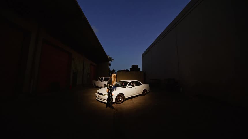 A woman gets off the hood of a white car parked in an alley between two buildings at dusk