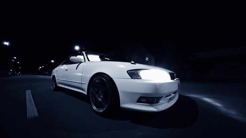 A white luxury automobile drives along a paved street at nighttime