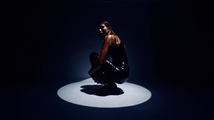 Woman crouches under a circular spotlight in a dark setting