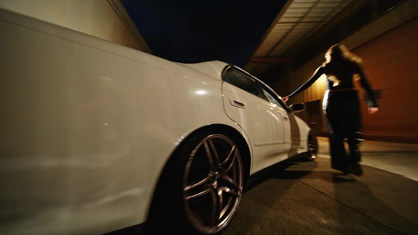 A view from behind a white car shows a person walking around while another vehicle sits nearby in a parking lot at night