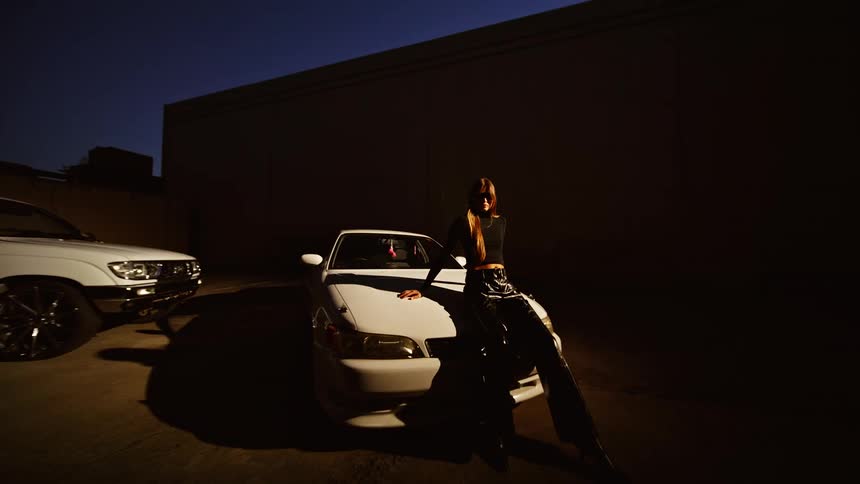 A woman in a brown jacket leans against the hood of a white car parked in a dark lot at night next to another white car and a tall building under a deep blue sky