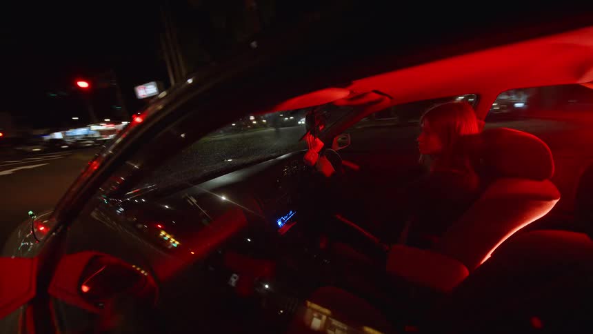 A view from the side shows a person sitting in the seat of a car while red lights glow from the inside as the vehicle drives down a dark street at night