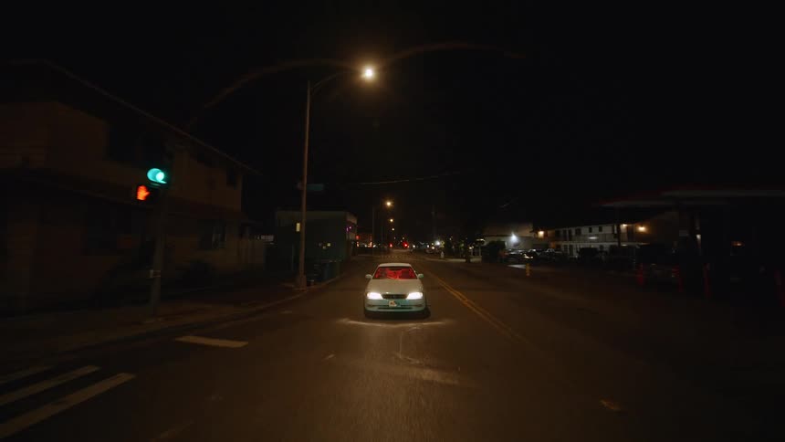 A view from the front shows a white car driving down a dark street at night while red lights glow from inside the vehicle