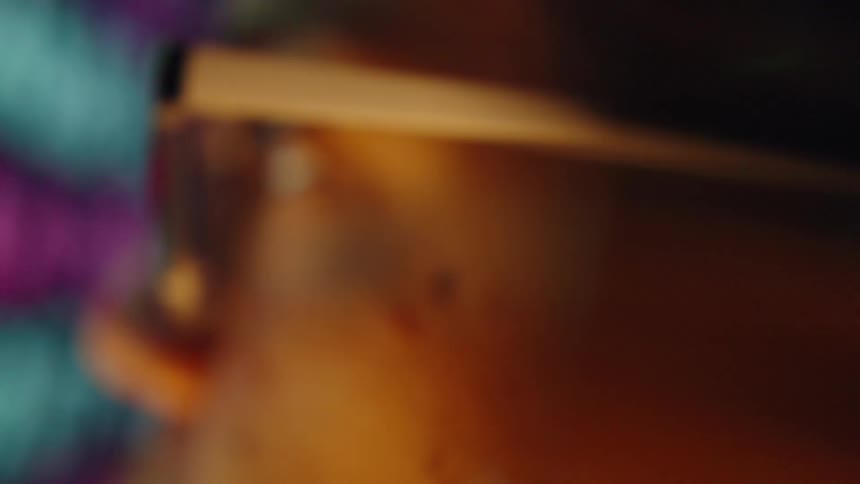 A warm heavily blurred closeup appears to show part of a persons face wearing glasses illuminated by amber lighting with colorful tones on a screen in the background