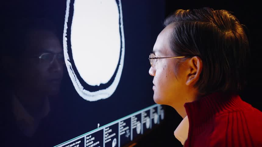 A man in a red sweater and glasses stares intently at a glowing medical monitorradiological scans of a human skull and brain cycle through the screen reflecting in the glass