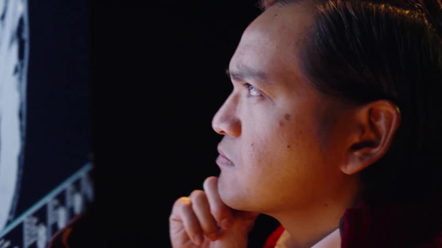 A male doctor in a red jacket rests his chin on his hand while intently studying a glowing brain mri scan on a large monitor in a dark room