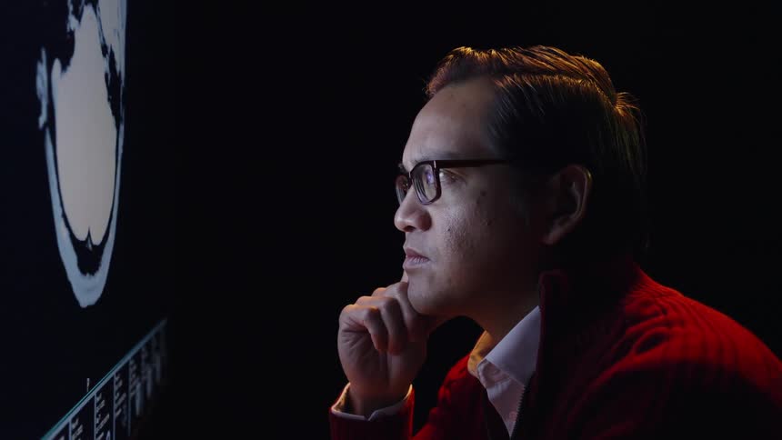 A man wearing glasses and a red sweater rests his chin on his hand as he studies a brightly lit screen in a dark room
