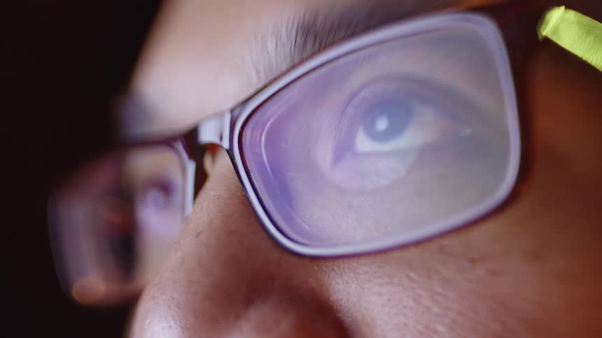 The video features a closeup shot of a persons eye behind glasses with the lenses reflecting blue and purple light from a screenthe person looks upward and moves their eyes slightly while the warm lighting emphasizes the details of their skin and t