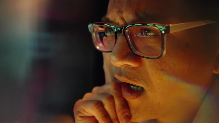A man wearing glasses looks intently at a glowing monitor in a dark room while resting his chin on his hand