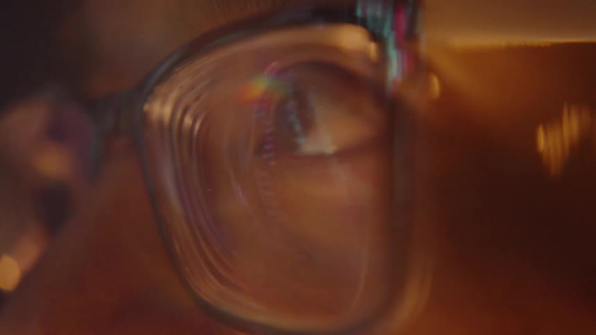 A man wearing glasses looks closely at a bright digital monitor which reflects colorful light and data onto the surface of his lens and into his eye within a dark room