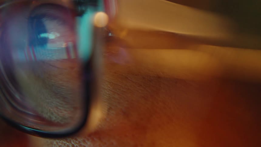The footage features an extreme closeup of a persons eye behind a pair of glasses with colorful light patterns and digital reflections dancing across the lensas the camera subtly shifts focus the warm ambient lighting highlights the texture of the