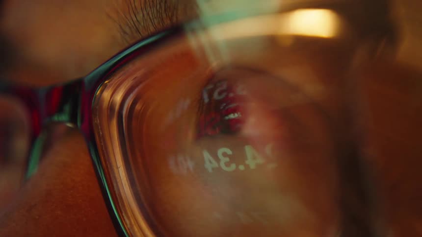 A close view of a man wearing glasses shows the reflection of scrolling green and red stock market data in his eyes while warm light glows in a dark room