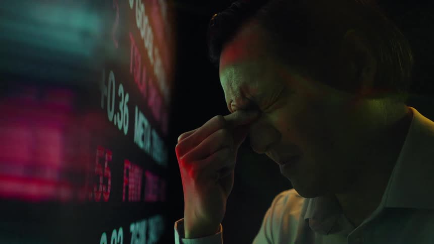 Man pressing the bridge of his nose in front of a board with financial quotes