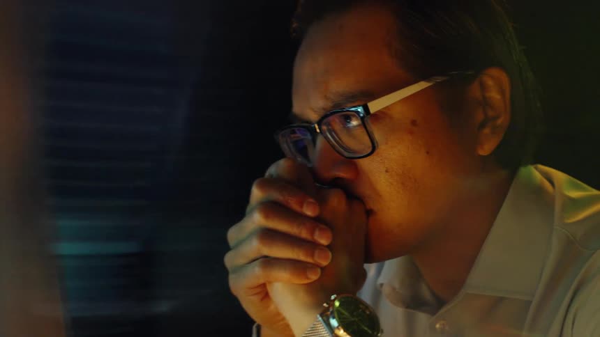 Young adult man wearing glasses and a white dress shirt looking at blurred screens indoors