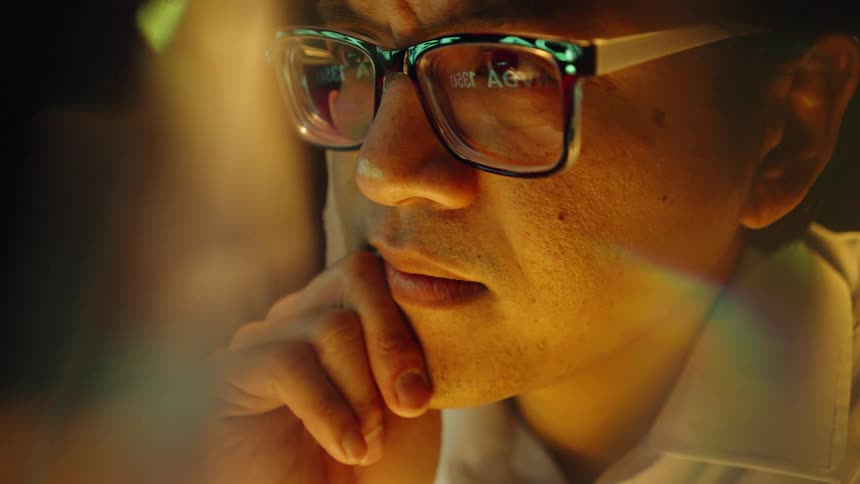 An asian man wearing glasses looks intently at a digital screen displaying stock market data and financial tickershis reflection is visible in the lenses of his glasses as he rests his chin on his hand with a focused contemplative expression